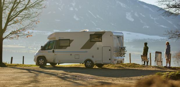 Photo by Sjoerd Verhelst on Unsplash een witte camperbus met twee mensen bij de oever van een meer in de bergen