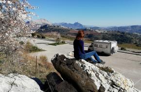 vrouw bij een bloesemboom bekijkt een camper in een berglandscha
