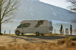 Photo by Sjoerd Verhelst on Unsplash een witte camperbus met twee mensen bij de oever van een meer in de bergen